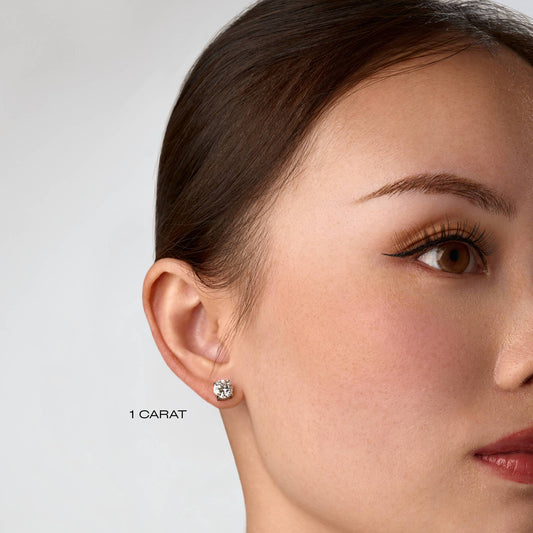 Close-up of a woman wearing a moissanite stone earring with '1 Carat' text on a plain background