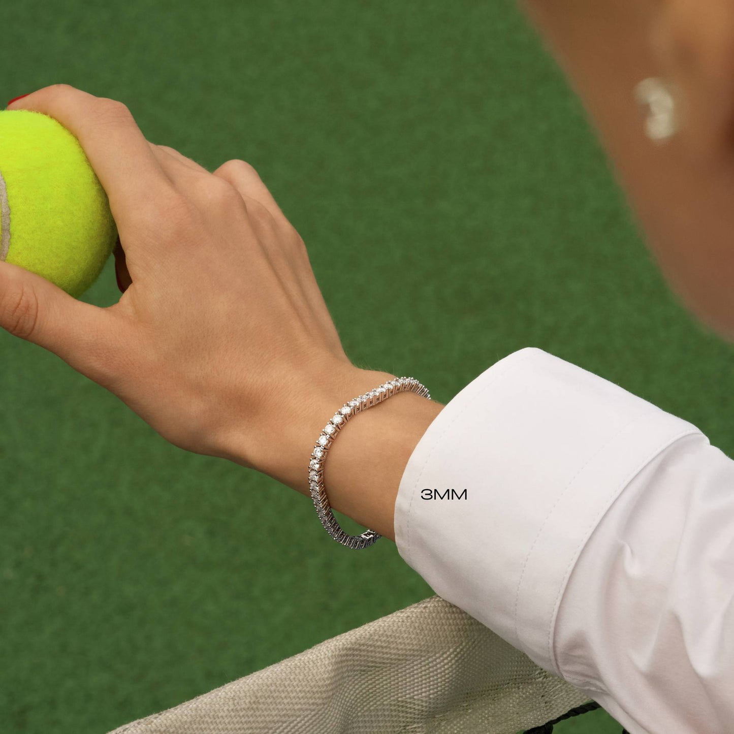 Hand holding a tennis ball with a tennis bracelet highlighting the width as 3mm on a green tennis court background