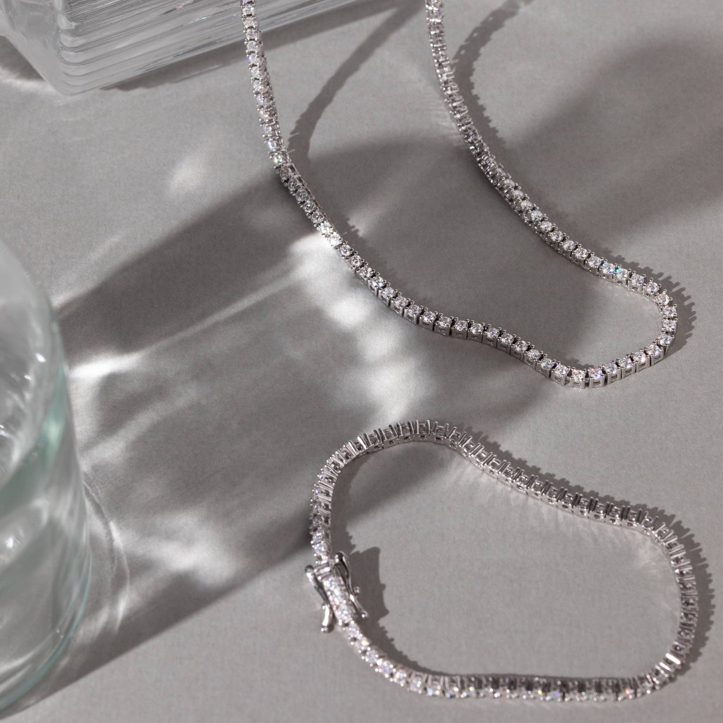 Silver chain tennis necklace on a light gray surface with a glass object in the corner.