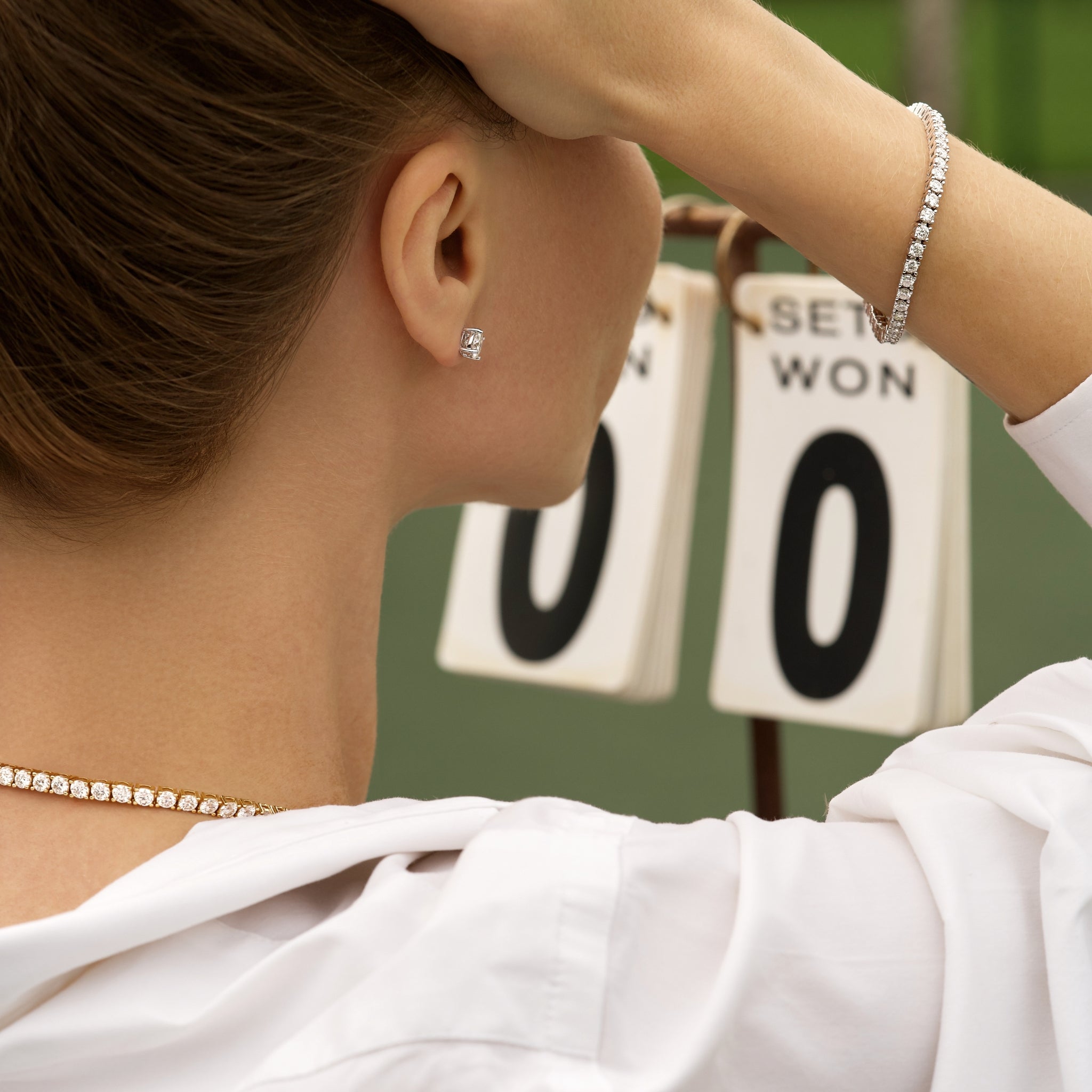 Woman wearing a moissanite earring, necklace and tennis bracelet with a tennis scoreboard in the background