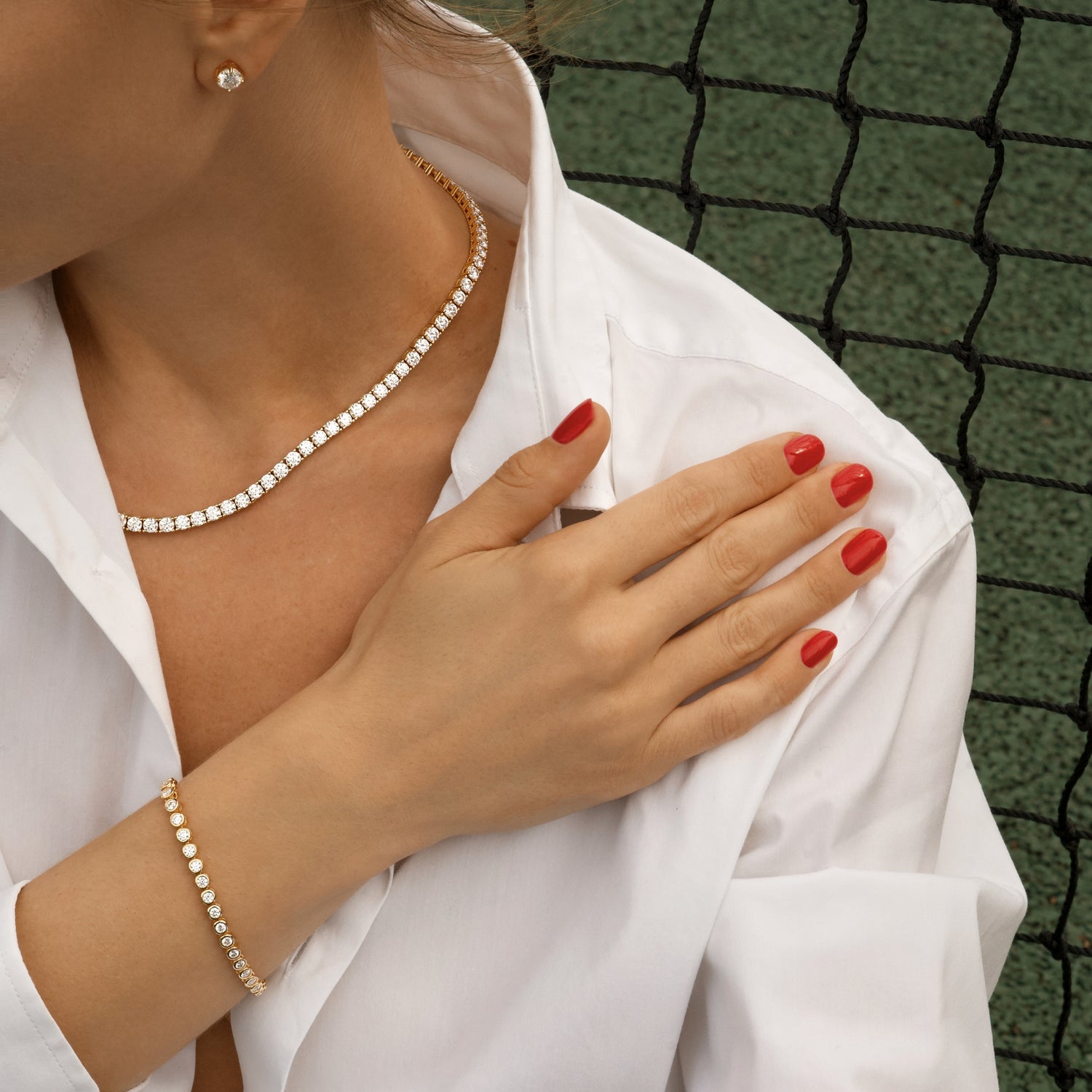 Person wearing a moissanite necklace, earrings and bracelet with red nail polish, standing against a tennis net.