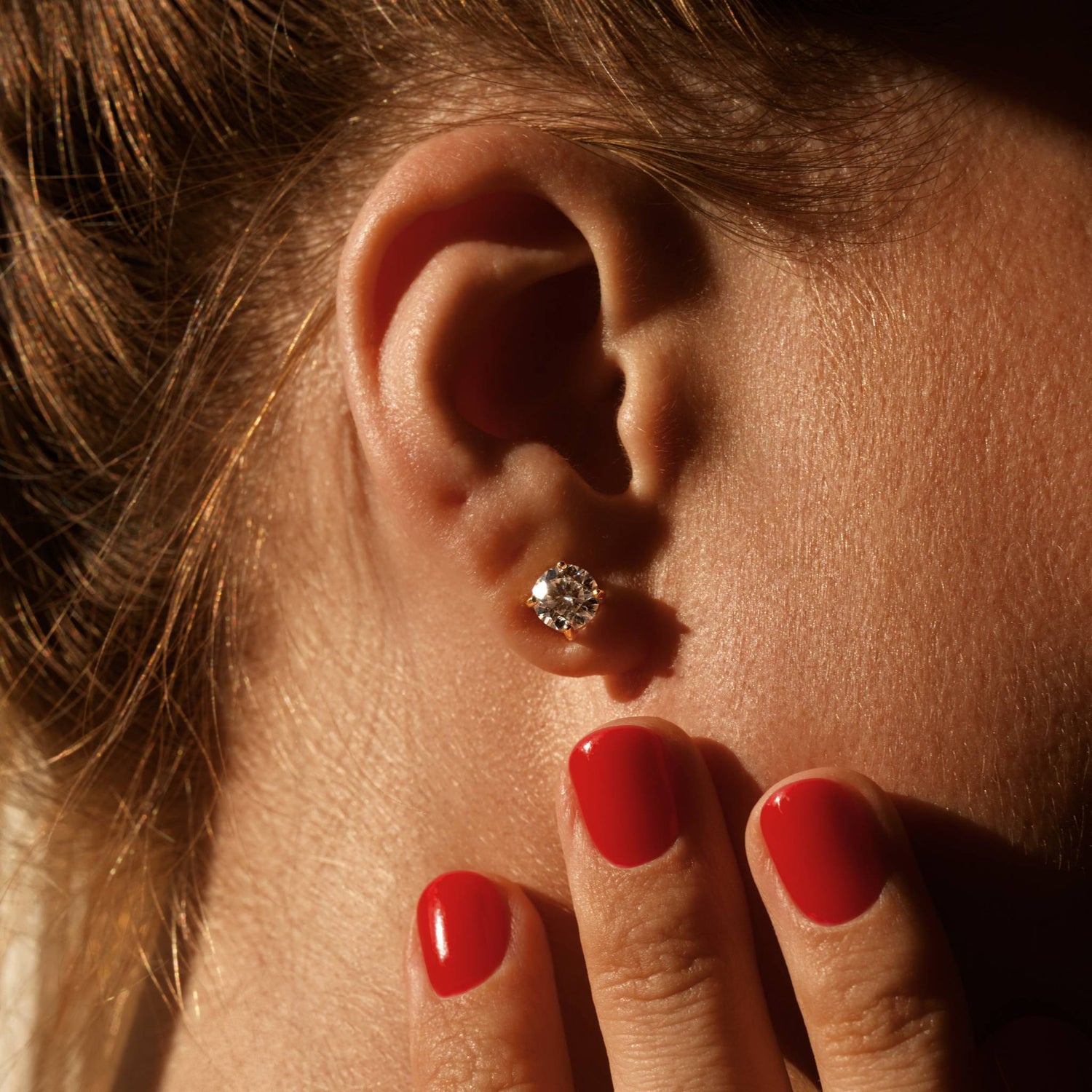 Close-up of an ear with a diamond earring, hand with red nails touching the earlobe.