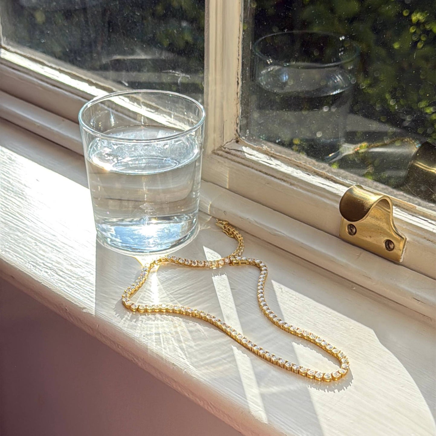 Glass of water and gold tennis necklace on a windowsill with sunlight