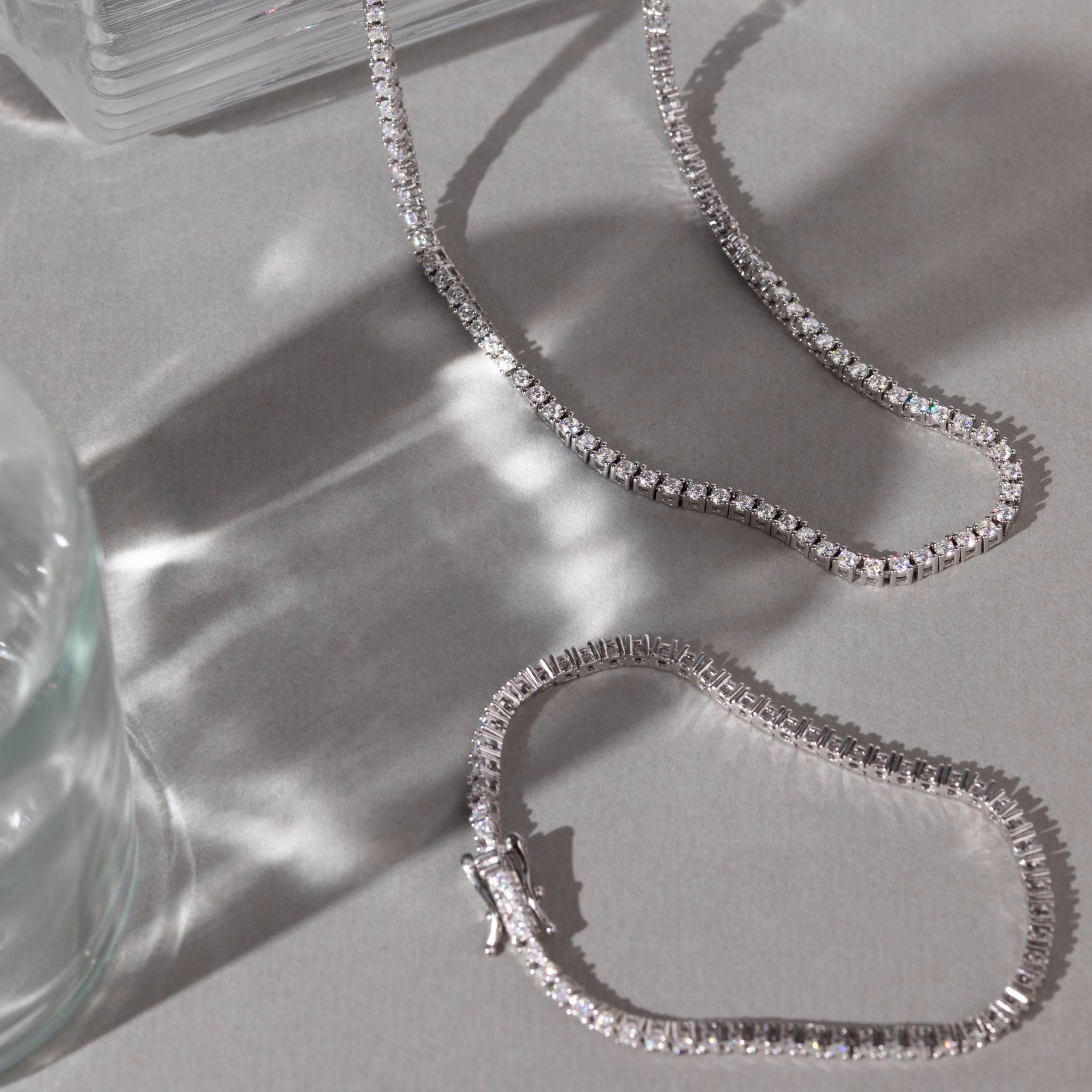 Silver chain tennis necklace on a light gray surface with a glass object in the corner.