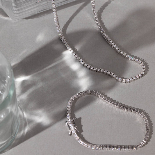 Silver chain tennis necklace on a light gray surface with a glass object in the corner.
