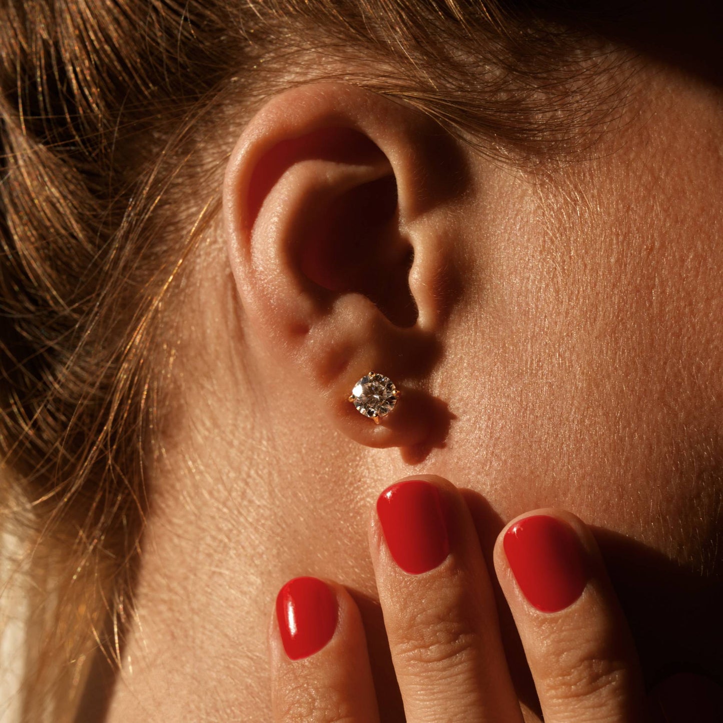 Close-up of an ear with a diamond earring, hand with red nails touching the earlobe.