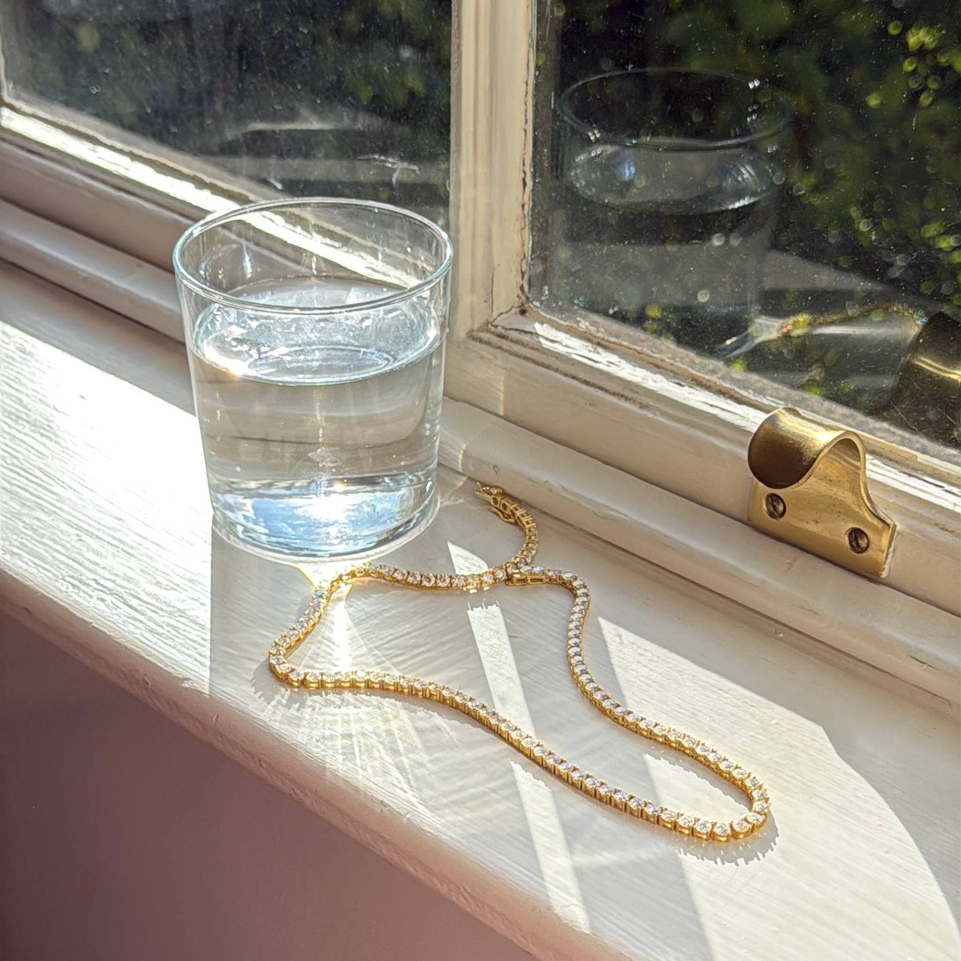 Glass of water and gold tennis necklace on a windowsill with sunlight